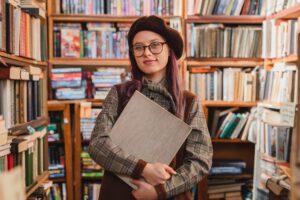 Portrait of an attractive cheerful 20s female hipster in glasses holding a book at bookstore