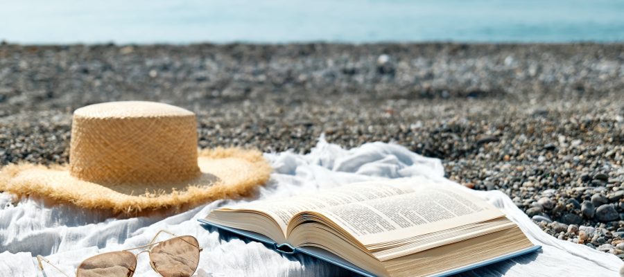 Open book, sunglasses and straw hat on towel on pebble beach. Concept of reading in summer vacation