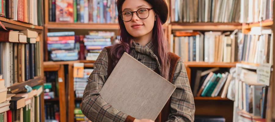 Portrait of an attractive cheerful 20s female hipster in glasses holding a book at bookstore
